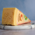 image of a large triangular wedge of Montasio cheese from Friuli region of Italy with the official montasio stamp visible in the cheese rind and the DOP markings on visible on the label, sat on a marble cheese board against a blue background
