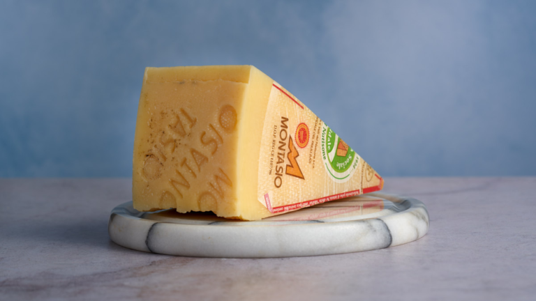 image of a large triangular wedge of Montasio cheese from Friuli region of Italy with the official montasio stamp visible in the cheese rind and the DOP markings on visible on the label, sat on a marble cheese board against a blue background