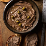 image of Crostini Neri in a black bowl dressed with capers on a wooden board surrounded by crostini spread with the pate