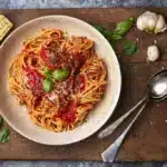 image of spaghetti alla Napoletana, a classic pasta dish of pasta with a vibrant tomato sauce from Naples served in a wide pasta bowl on a wooden board, surrounded by tomatoes, parmesan, garlic and basil