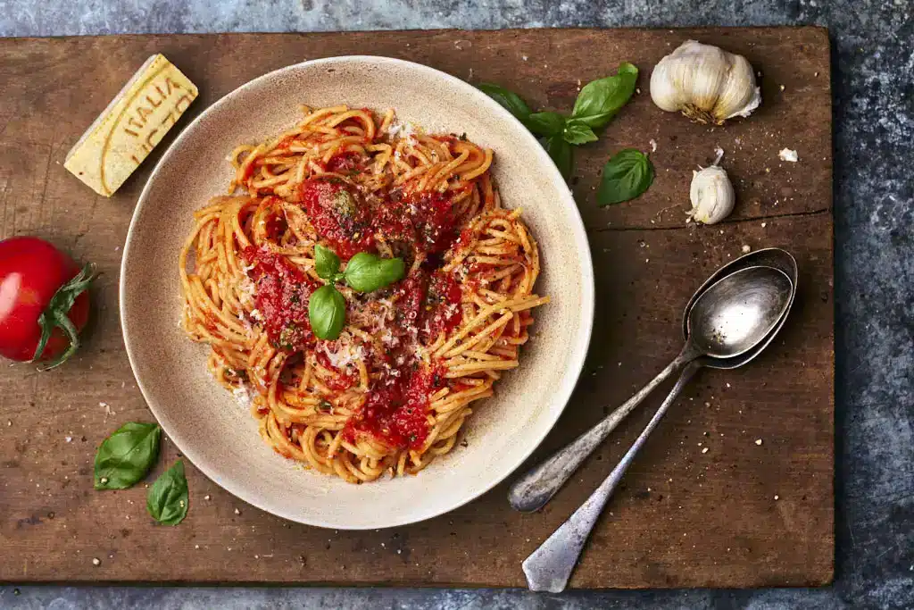 image of spaghetti alla Napoletana, a classic pasta dish of pasta with a vibrant tomato sauce from Naples served in a wide pasta bowl on a wooden board, surrounded by tomatoes, parmesan, garlic and basil