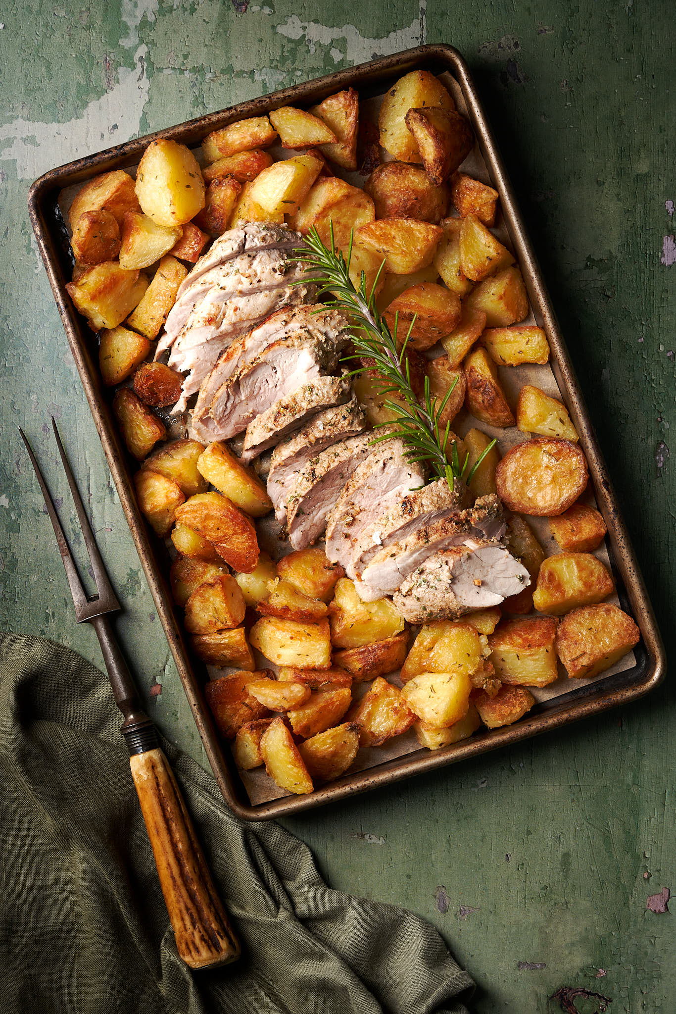 I'm age of Friulian loin of pork with roast potatoes served in a tray on a green background with a dark green background and carving fork