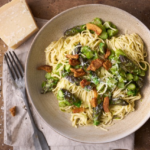 image of taglierini con asparagi e guanciale, a central italian dish of thin angel hair pasta with asparagus and pork, served in a wide pasta bowl on a wooden board, with a block of parmesan cheese on the side.