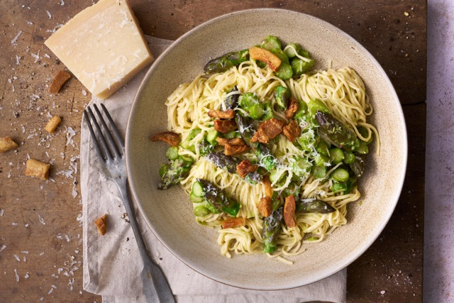 image of taglierini con asparagi e guanciale, a central italian dish of thin angel hair pasta with asparagus and pork, served in a wide pasta bowl on a wooden board, with a block of parmesan cheese on the side.