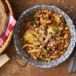 image of trippa alla genovese, a tripe dish from Liguria made with tripe, pine nuts and mushrooms in a tomato sauce served in a rustic pan on a wooden board with some crusty bread
