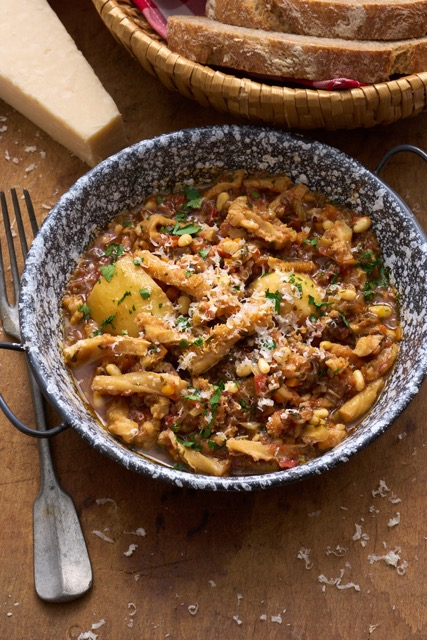 image of trippa alla genovese, a tripe dish from Liguria made with tripe, pine nuts and mushrooms in a tomato sauce served in a rustic pan on a wooden board with some crusty bread