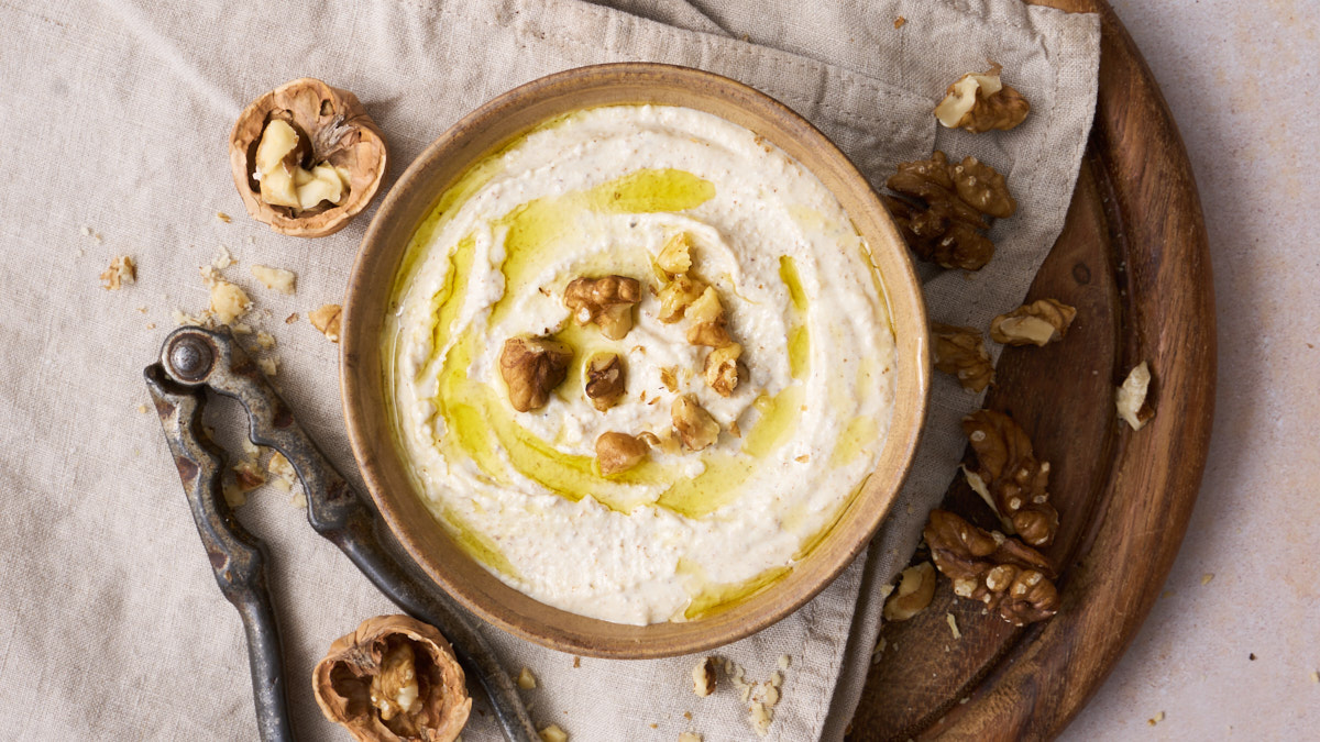 image of salsa di noci, the ligurian walnut sauce served in an earth coloured bowl, drizzled with golden olive oil and garnished with walnut pieces, presented on a wooden board with a nut cracker and cracked walnut shells and walnut pieces on the board.