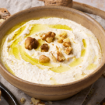 image of salsa di noci, the ligurian walnut sauce served in an earth coloured bowl, drizzled with golden olive oil and garnished with walnut pieces, presented on a wooden board with a nut cracker and cracked walnut shells and walnut pieces on the board.