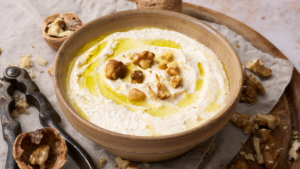 image of salsa di noci, the ligurian walnut sauce served in an earth coloured bowl, drizzled with golden olive oil and garnished with walnut pieces, presented on a wooden board with a nut cracker and cracked walnut shells and walnut pieces on the board.