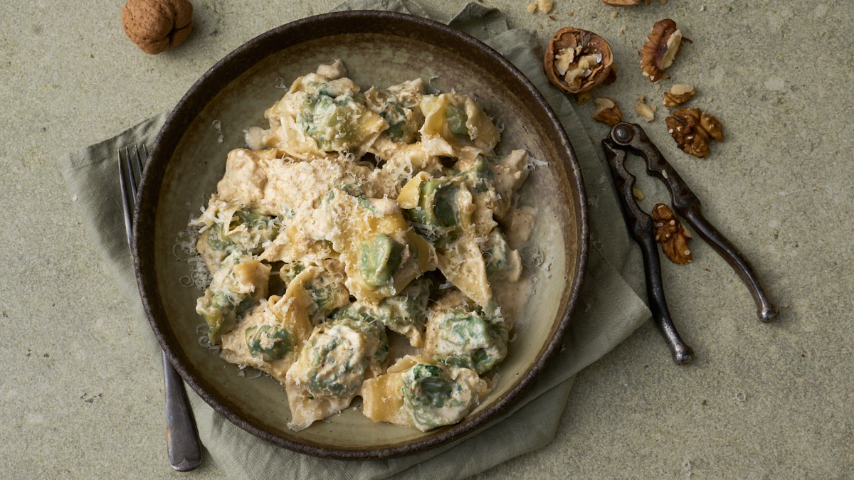 image of pansoti con salsa di noci, a Ligurian filled pasta dish of greens dressed in a walnut sauce, served in wide bowl surrounded by whole and broken walnuts and walnut pieces.
