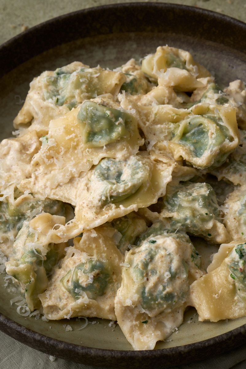 image of pansoti con salsa di noci, a Ligurian filled pasta dish of greens dressed in a walnut sauce, served in wide bowl surrounded by whole and broken walnuts and walnut pieces.