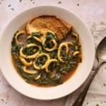 landscape image of calamari in zimino, a Ligruian stew of calamari with chard, served in a wide off white bowl with a slice of rustic toast in teh bowl soaking up all the juices.
