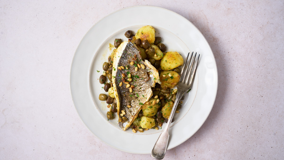image of orate alla ligure - a ligurian sea bass dish served with potatoes, pine nuts and Taggiasca olives, served on a white plate with a clean background