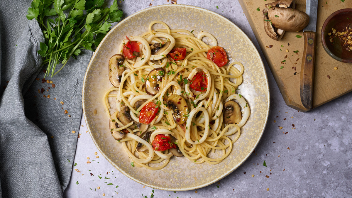 image of spaghetti mare e monti, a pasta dish with calamari rings and mushrooms, served on a wide plate next to a rustic wooden board with a wooden handled knife and some raw mushrooms. from the Marche region