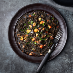 image of tagliolini al nero di seppia con trota ‘fil di fumo’, black spaghetti served with San Daniele smoked trout on a black plate with a dark grey background