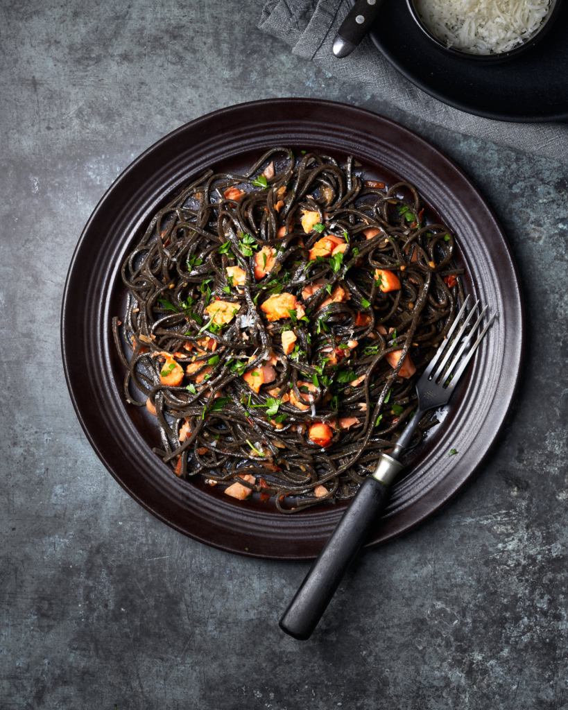 image of tagliolini al nero di seppia con trota ‘fil di fumo’, black spaghetti served with San Daniele smoked trout on a black plate with a dark grey background