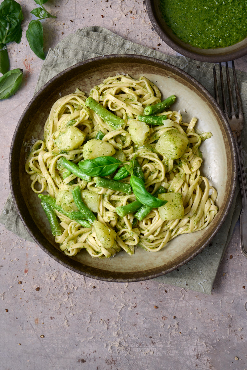 portrait image of trennete al pesto, a dish of long trennete pasta dressed in pesto with potatoes and green beans from the Liguria region of Italy, served in a wide rustic bowl surround by a bowl of pesto and basil leaves.
