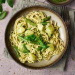 portrait image of trennete al pesto, a dish of long trennete pasta dressed in pesto with potatoes and green beans from the Liguria region of Italy, served in a wide rustic bowl surround by a bowl of pesto and basil leaves.