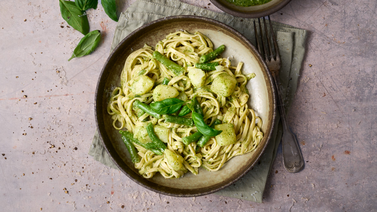 portrait image of trennete al pesto, a dish of long trennete pasta dressed in pesto with potatoes and green beans from the Liguria region of Italy, served in a wide rustic bowl surround by a bowl of pesto and basil leaves.