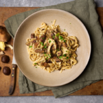 image of pasta con castagne e funghi, an italian autumn dish with chestnuts and woodland mushrooms in a rustic bowl set on a green napkin on a wooden board with raw mushrooms and chestnuts in the background