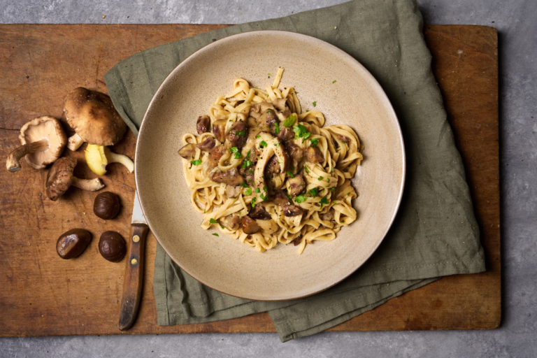 image of pasta con castagne e funghi, an italian autumn dish with chestnuts and woodland mushrooms in a rustic bowl set on a green napkin on a wooden board with raw mushrooms and chestnuts in the background